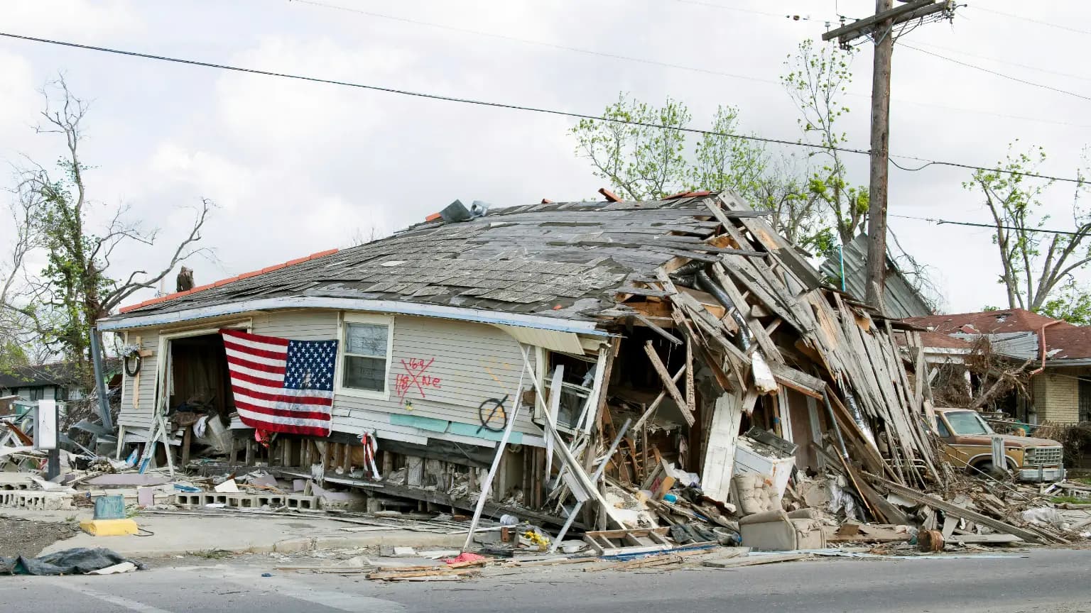 A partially collapsed house, surrounded by debris, with an American flag hanging from the damaged exterior.